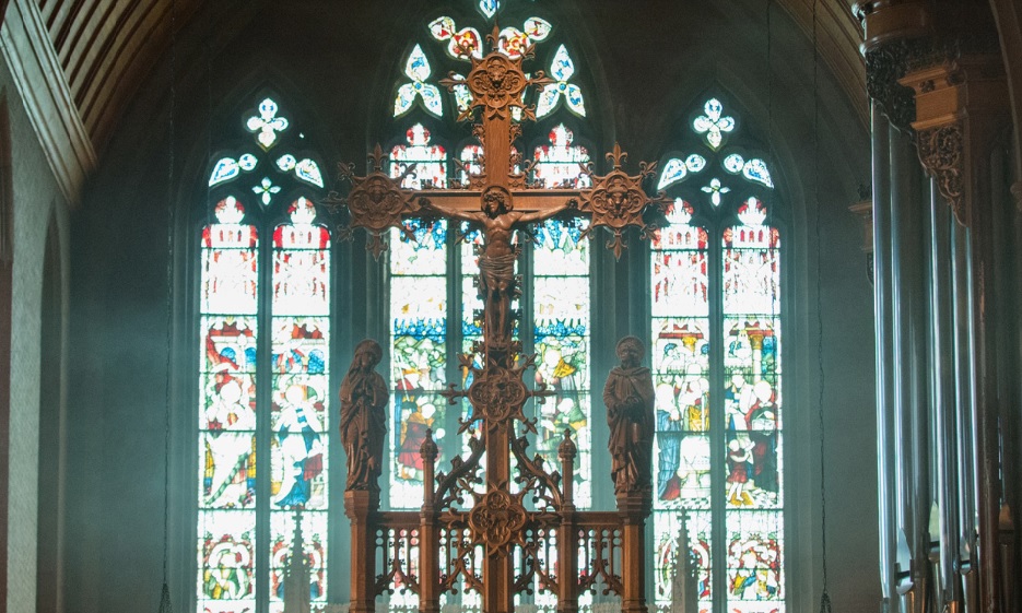 A wooden crucifix in front of three stained glass windows.