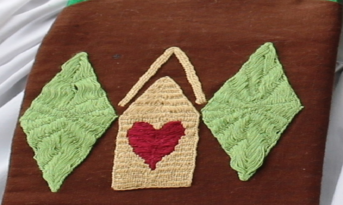 Embroidery of a yellow house with a red heart between two light green diamonds on a brown background.